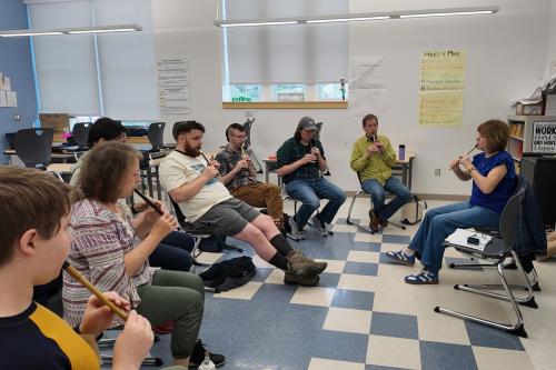 Seven people of all ages play fife, seated in a row in a brightly lit classroom, facing a teacher also playing.
