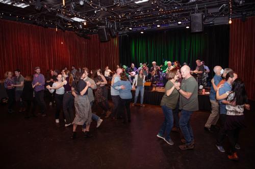 Several pairs of people dance in a music venue while a band plays. One person on the floor, wearing a headset mic, gives directions.
