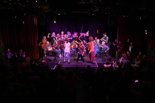 Musicians and dancers under purple lights on a small stage, surrounded by a crowd.
