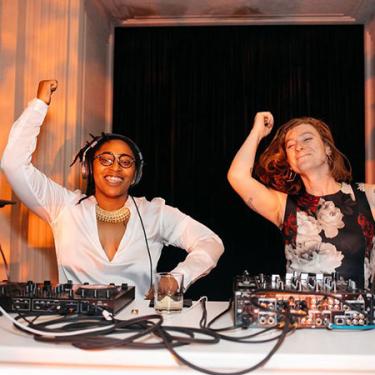 Two women at a DJ booth, dancing in unison.