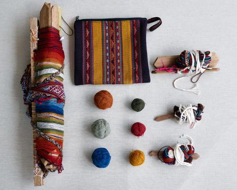 A small woven handbag, six balls of dyed yarn, three small weaving tools, and one larger loom with a striped woven patterns already started, rolled up.