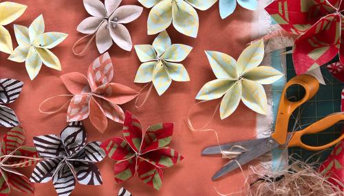 Closeup on a pair of hands folder paper into the petals of a flower. The table is scattered with yellow and pink paper flowers and craft supplies.
