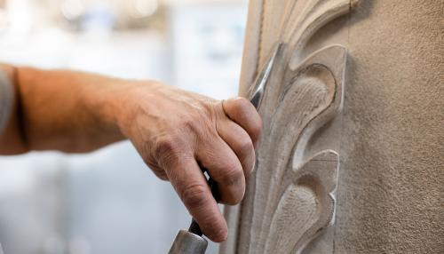 Closeup of a hand holding a chisel, pointed upwards and ready to carve into a piece of white, ornate stone on the right.