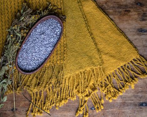 Pieces of yellow woven textile, a small bowl of blueish powder, and sprigs of a plant with yellow flowers.