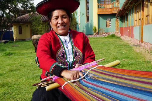A woman smiles, weaving outdoors on a loom secured around her waist.
