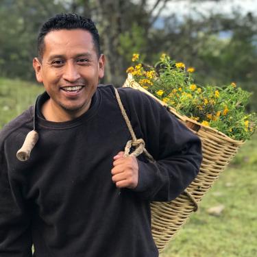 A man smiles, carrying a large basket full of yellow flowers over his shoulder.