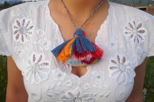Closeup of a woman’s chest; around her  neck is a large yarn tassel on a necklace.