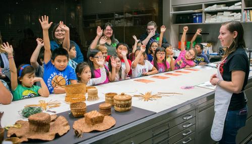 A crowd of young school children and a few adult chaperones surround one side of a work table, each raising one hand. A woman in an apron stands on the other side of the table smiling. On the table are several baskets and objects made of woven plant mater