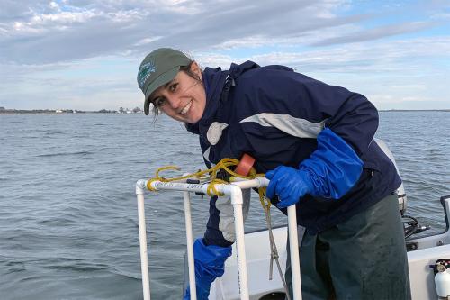 A woman in waterproof jacket, pants, and big sloves smiles aboard a boat in a bay.