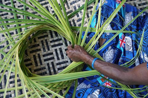 Overhead closeup of a person sitting cross-legged on a woven straw mat, weaving a green palm leaf  into a circular shape.