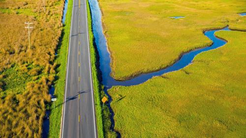 Aerial photo in saturated colors, showing a two-lane road cutting through yellow and green wetlands, intersected by vibrant blue waterways.