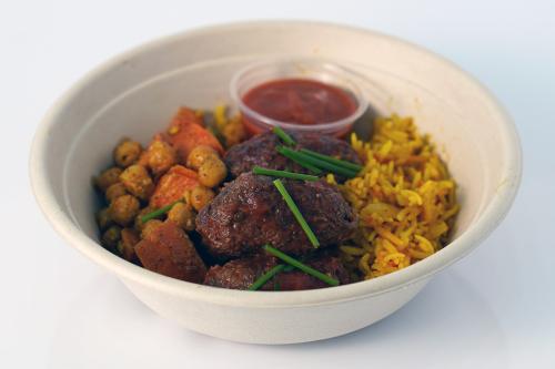 A cardboard bowl full of chunks of brown meat, yellow rice, chickpeas, and orange carrots, with a small cup of red sauce.
