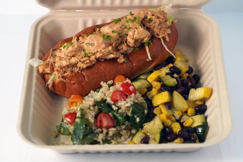 Hoagie-style sandwich filled with chunks of fish and shredded cabbage in an orange sauce and garnished with green chives, next to grain and vegetable salads.