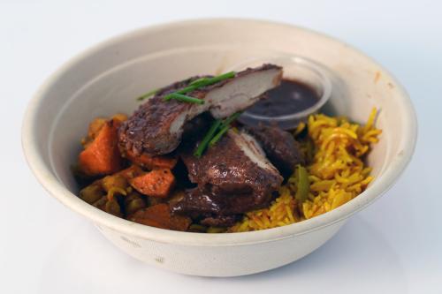 A bowl with pieces of chicken in dark brown sauce, yellow rice, orange carrots, and cup of dark brown sauce.
