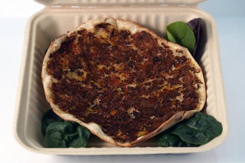 Round flatbread with reddish brown meat topping, set in a to-go box with a few pieces of green lettuce.