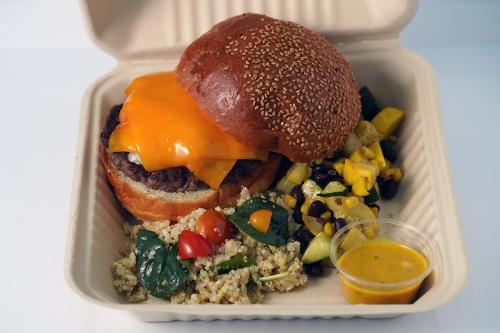 Burger with melty orange cheese and sesame seed bun, alongside grain and vegetable salads, with a cup of yellow dressing. 