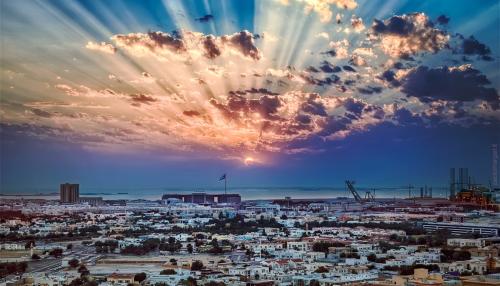 Wide shot of city at sunset, sun rays visible through scattered clouds.