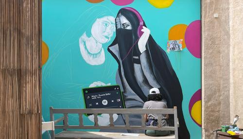 A person seated, facing away, paints a large mural depicting an Arab woman wearing head and face coverings, listening to pink headphones connected to a device playing music, on a background of bright teal with pink and orange dots.