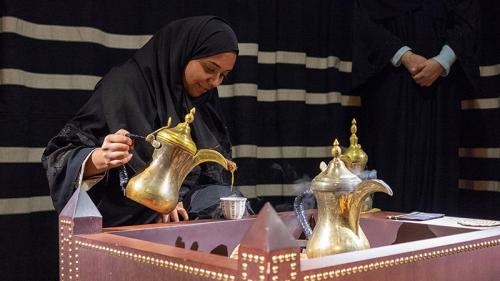A woman in a black abaya (robe), her face just out of frame, pours coffee from a shiny, golden samovar into a small patterned cup. Two other steaming savors await use.