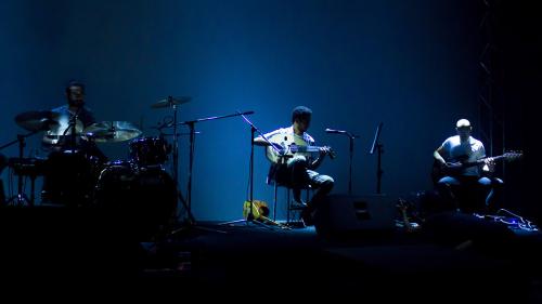 Three-person band on a dark stage, barely lit blue. They play drums, oud, and electric bass.