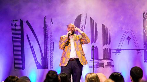 A person speaking on stage for a crowd of people against a purple-lit backdrop.