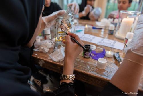 A person in a hijab, facing away, paints a clear glass jar with a black and gold design. Others around a table appear to be doing the same.