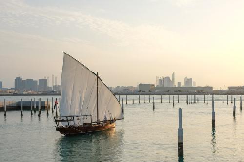 A small wooden boat with a white mast floats in a harbor, with a city skyline in the distance.