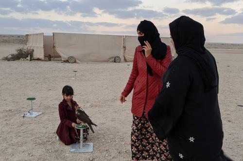 Two women stand in a vast desert landscape, watching a girl kneeling in the sand attending to a falcon on a perch.