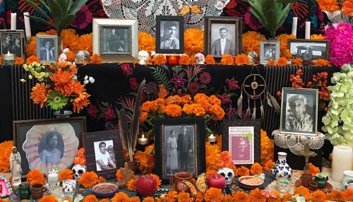 Altar set with framed black-and-white photos of people, orange marigolds, white candles and skull figurines, and other objects.