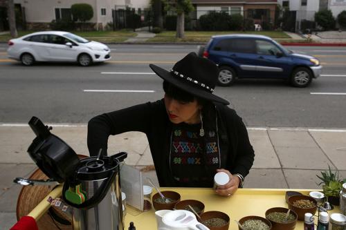 A person in black hat and jacket sits outdoors, cars passing by on the street in the background, arranging containers of seeds on a flat surface.