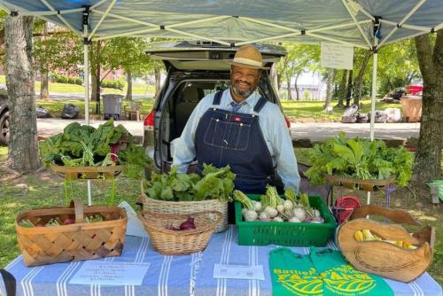 A man in hat and overalls stands behind a table, full of baskets of produce, under a tent.