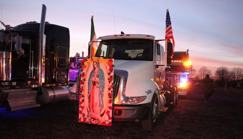 Parked semi truck cabs at dusk. The one in front has a painting of the Virgin Mary, adorned with red and yellow flowers, on the hood.