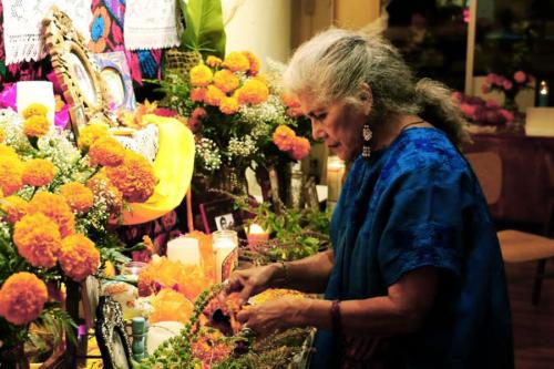 A woman with gray hair pulled back adjusts the pieces of an altar, including photos and orange marigold flowers.