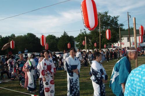 A crowd of people line up on a grassy field, under red and white lanterns, wearing long kimonos.