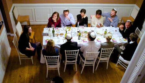Twelve people sit around a rectangular dinner table set with glasses, wine bottles, and pamphlets. The men wear white yamulkes.