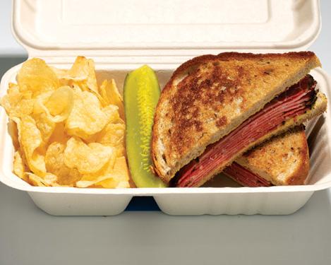 A sandwich with sliced pink pastrami in a cardboard contained with potato chips and a green pickle slice.