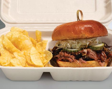 Pulled  pork on a bun with green pickles and white slaw with potato chips in a cardboard container. 