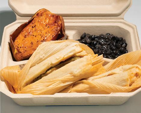 Two tamales wrapped in corn husks, with orange sweet potatos and black beans in a cardboard container.