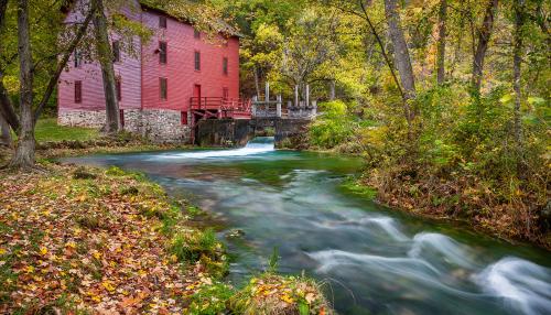 Three-story red brick building along the  banks of a river through the woods.