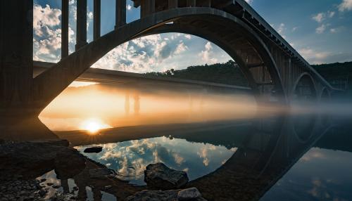 Reflection of the sunrise on the water, underneat an arched bridge.