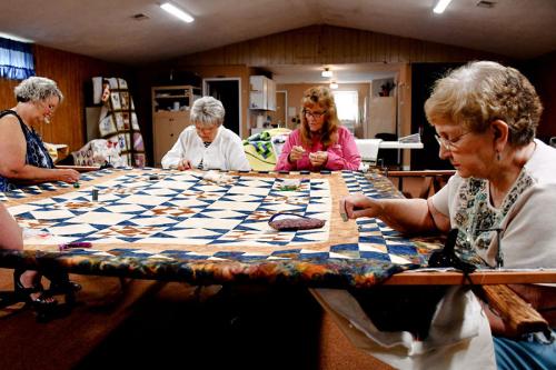 Four women sit at a table, working togethered on a patterened quilt.