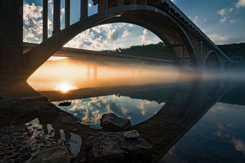 Reflection of the sunrise on the water, underneath an arched bridge.