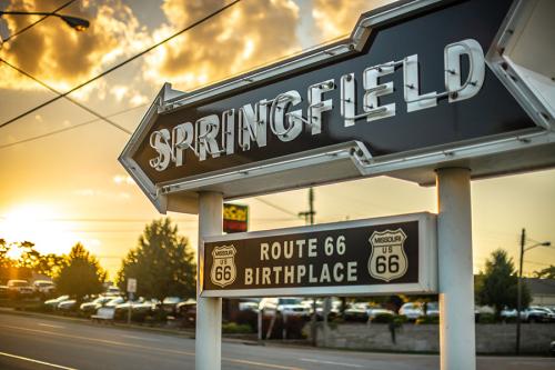 Neon road sign shaped like an arrow, with the name Springfield. Under it, a sign for Route 66 Birthplace. Brilliant sunset in the background.
