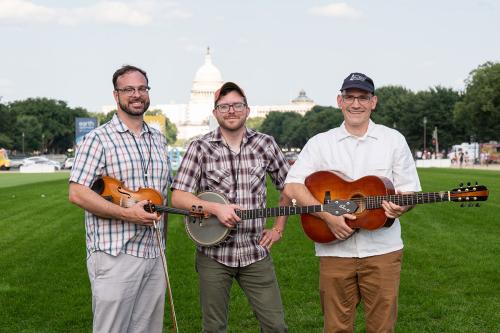 Three men pose in front of the U.S. Capitol Building, each holding a musical instrument: fiddle, banjo, and acoustic guitar.