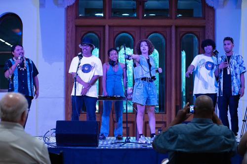 Several musicians stand on stage indoors under blue light.
