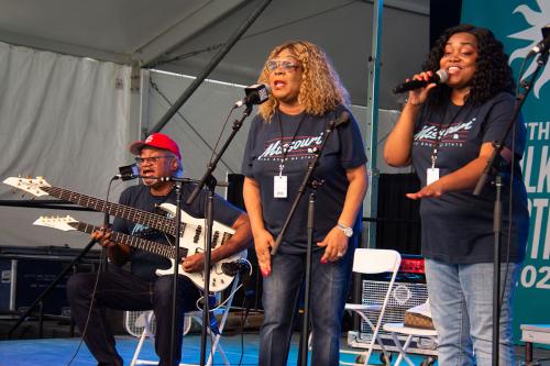 Two women sing and a man plays double-necked guitar on stage.