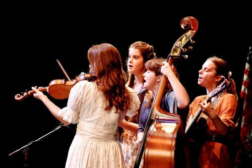 Four young musicians performing on stage with fiddle, upright bass, and acoustic guitar.