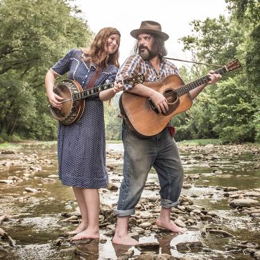 Man and woman playing banjo and guitar in a shallow creek.