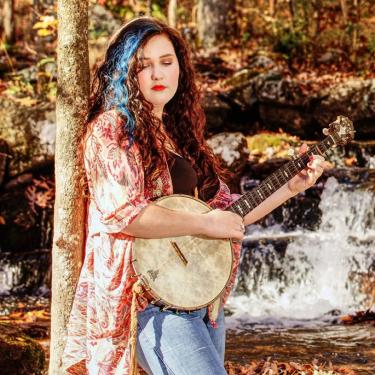 Grace Stormont holding banjo with small waterfall in the background.