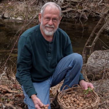 A man kneels in leaves in front of a river with a basket of nuts.